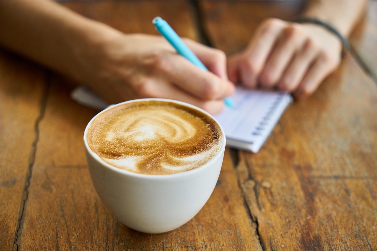 A perfectly crafted cappuccino with latte art sitting on a wooden table next to a fresh croissant in a warm, sunlit coffee shop